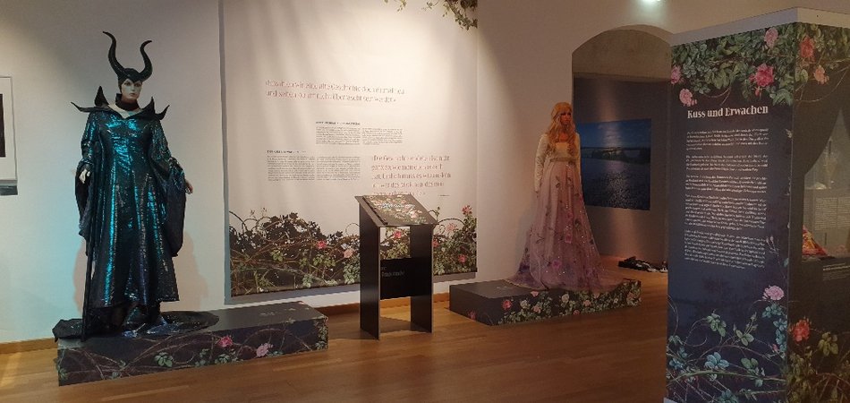 Der Blick in die Ausstellung zeigt auf zwei mit Rosenhecken bebilderten Podesten stehende lebensgroßen Figuren des Dornröschen und der Bösen Königin, an der Wand dahinter befindet sich ein Ausstellungstext