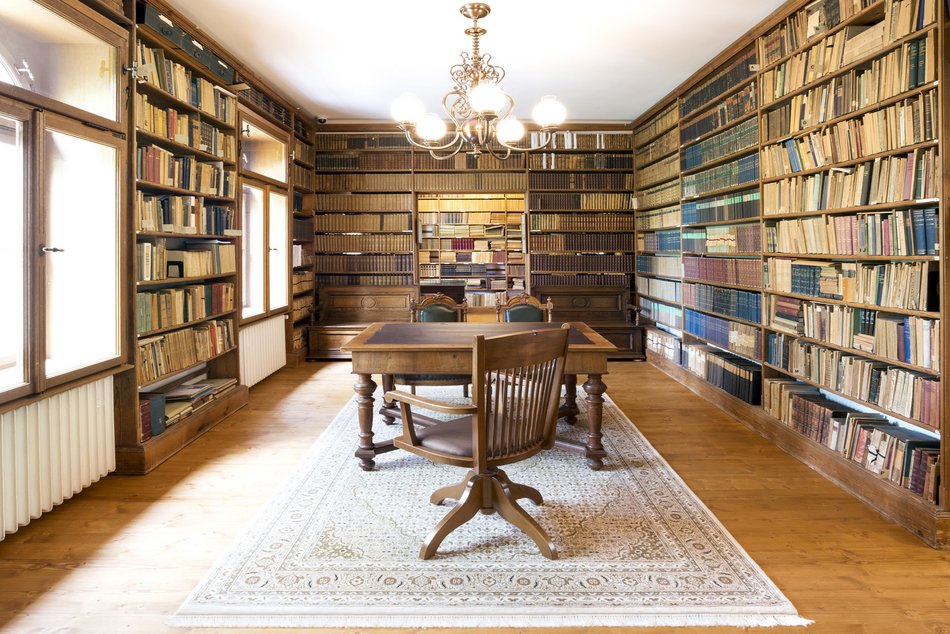 Blick in eine historische Bibliothek, deren prall gefüllte Massivholz-Bücherregale drei Wände einnehmen. In der Mitte ein großer, alter Holztisch mit drei Stühlen.