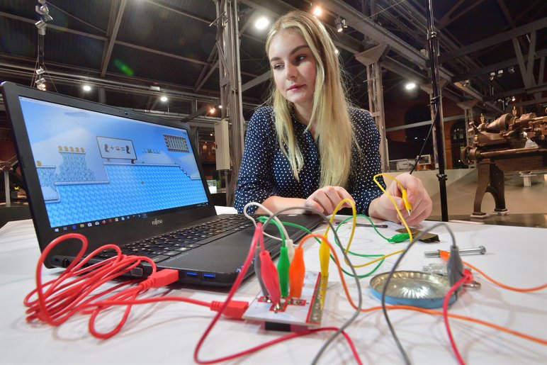 Eine junge Frau sitzt an einem Werktisch und schaut auf einen Bildschirm, während sie mit dem Ende eines Kabels einen Stromkreis zu schließen scheint. Mit dem Laptop verbunden ist ein rotes Kabel, weitere Kabel in Orange, Gelb, Grün und Weiß komplettieren den Stromkreis.