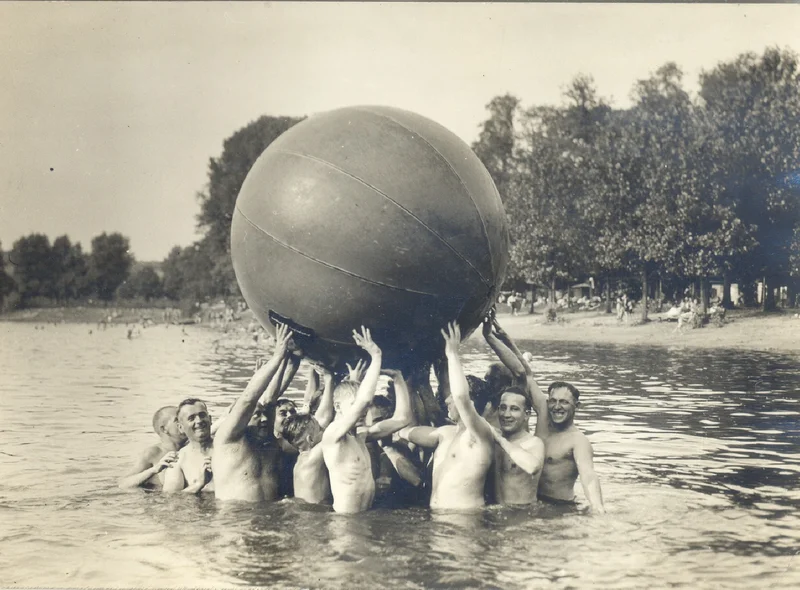 Historische Schwarzweißfotografie mehrerer badender Männer, die einen riesigen Wasserball in die Höhe halten