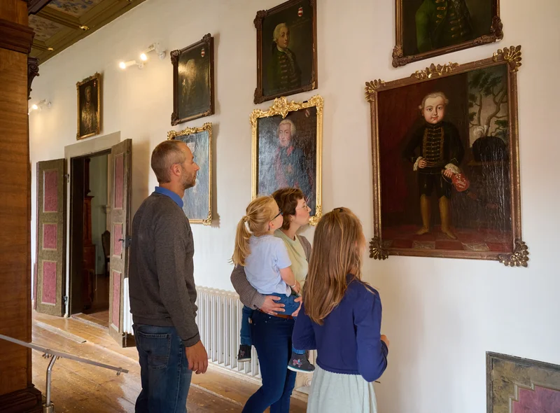 Eine Familie betrachtet goldgerahmte Gemälde an der Wand. Auf einem Bild ist ein Kind in historischer Kleidung zu erkennen.