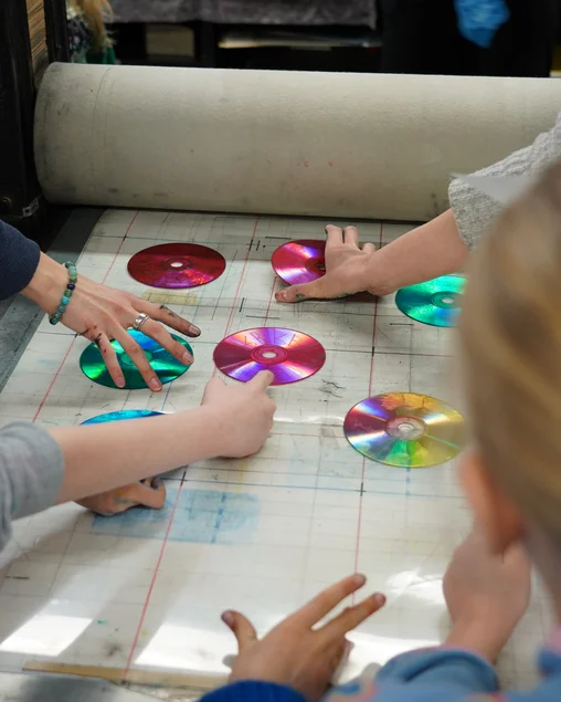 Tiefdruck mit CDs Mehrere Kinderhände arrangieren CDs, in welche Bilder eingeritzt sind, vor einer Druckerpresse.