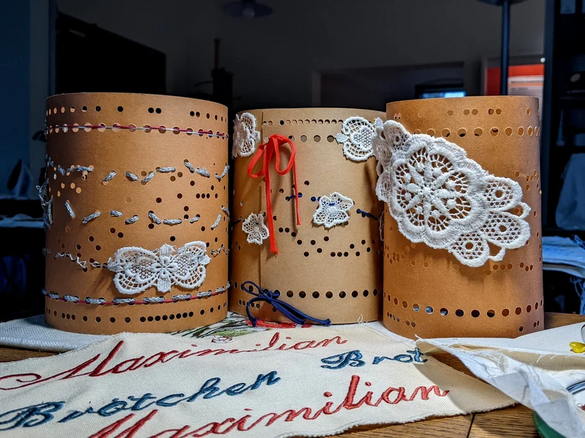 Gestalten mit Spitze Drei historische Lochkarten sind mit Beispielen für Gestaltungen mit Spitze versehen, davor liegt ein Tuch mit der gestickten Aufschrift „Maximilian – Brot – Brötchen“.