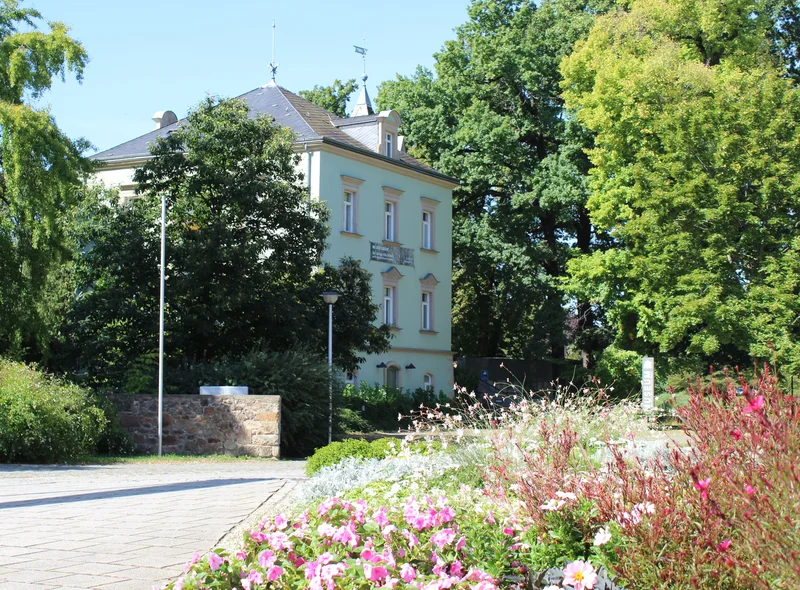 Die Karrasburg in Coswig, ein dreistöckiges, lindgrünes Gründerzeitgebäude umgeben von großen, grünen Laubbäumen
