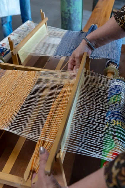 Museumspädagogik: Weben Nahaufnahme eines Miniatur-Webstuhls, in den Garne eingespannt sind. Es ist zu sehen, wie zwei Hände ein Schiffchen mit Garn durch die gespannten Fäden führt.
