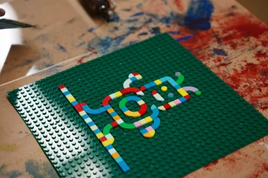 Auf einem mit Farbflecken übersäten Tisch liegt eine grüne Lego-Grundplatte, auf der bunte Lego-Steine ein abstraktes Muster bilden.