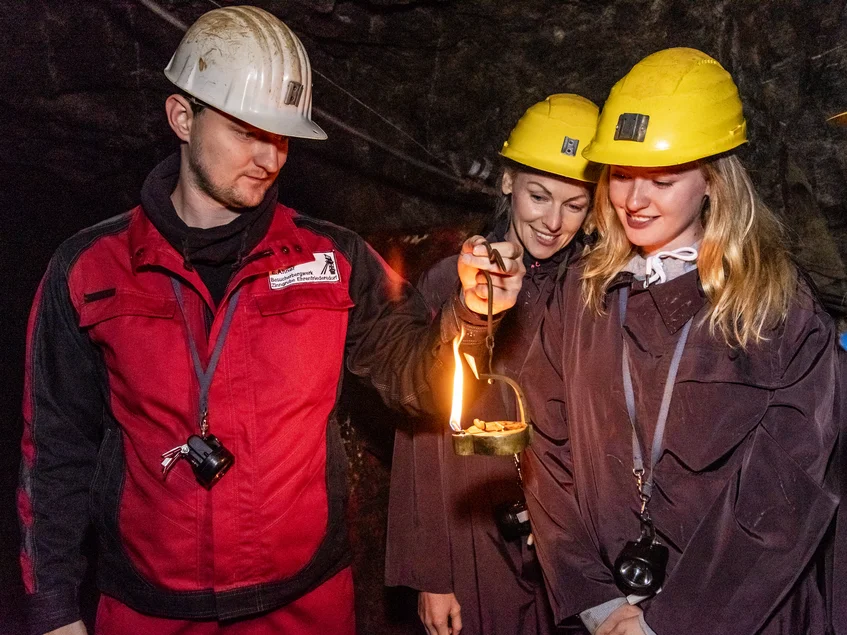 Im Besucherbergwerk In einem Stollen stehen nebeneinander ein Mitarbeiter der Zinngrube in Schutzkleidung (Schutzjacke, Schutzhelm) und zwei Besucherinnen in Schutzkleidung (braune Overalls, Schutzhelme). Der Mitarbeiter hält eine angezündete, mittelalterliche Froschlampe hoch, auf die beide Besucherinnen erstaunt blicken.