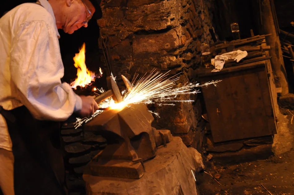 Schmied im Frohnauer Hammer Das Foto zeigt einen nach historischem Vorbild gekleideten Schmied (einen „Hammergesellen“) im Frohnauer Hammer, mit einem Hammer bearbeitet er ein Stück glühendes Eisen, sodass die Funken fliegen.