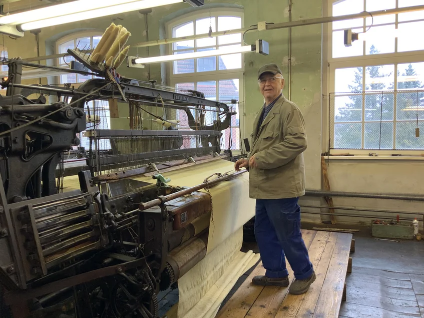 Museumsmitarbeiter am Webstuhl Ein älterer Mann in Arbeitskleidung steht an einer großen historischen Industriewebmaschine.