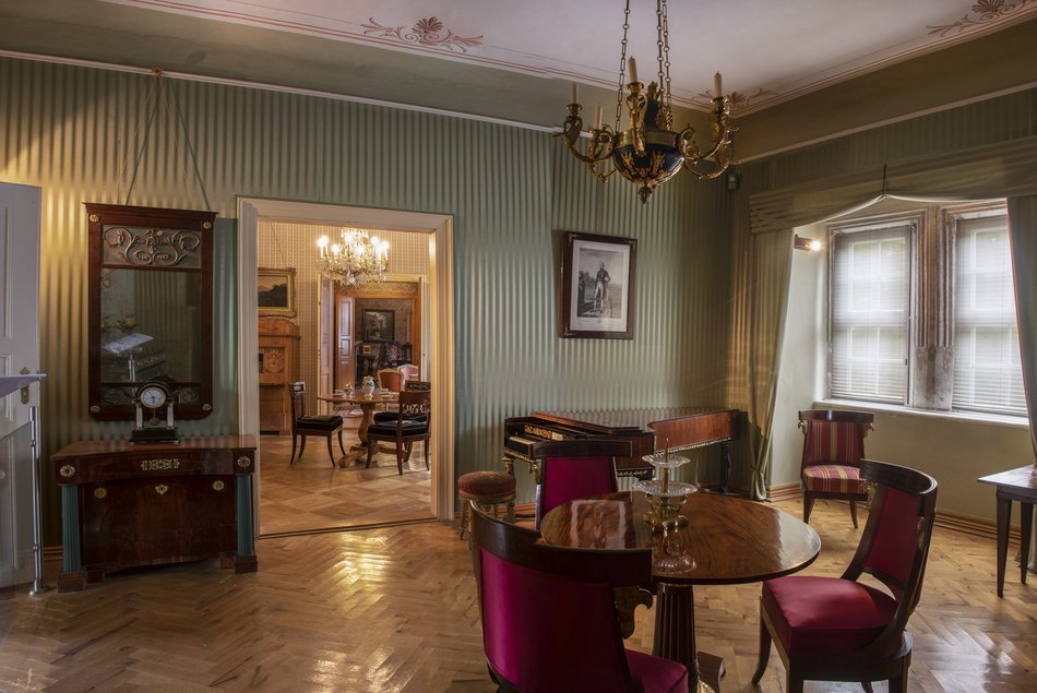Foto eines mit Originalmöbeln im Empire-Stil ausgestatteten Ausstellungsraums im historischen Schloss Hinterglauchau. Der Blick richtet sich auf den Durchgang zum nächsten Zimmer, welches ebenfalls historische Möbel enthält. An den Decken hängen Kronleuchter, die Böden sind mit Parkett ausgelegt.