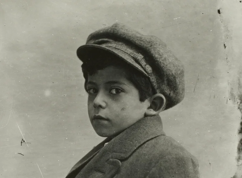 Historisches Porträtfoto eines Jungen mit Hut, der direkt in die Kamera schaut. Das Foto ist in Schwarzweiß.