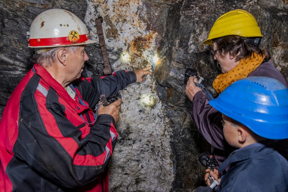 In einem Stollen stehen drei Personen vor einer natürlichen Steinwand. Links ein Mitarbeiter der Zinngrube in Schutzkleidung (Schutzjacke und Schutzhelm) und rechts zwei Besucher:innen in Schutzkleidung (Overalls und Schutzhelme, eine erwachsene Frau und ein Kind). Alle drei Personen halten kleine Taschenlampen in der Hand und leuchten auf einen Punkt an der Wand mit hellem Gestein, auf den der Mitarbeiter deutet.