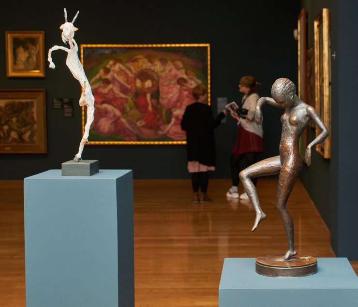 Im Raum stehen zwei sich zugewandte Frauen, an den Wänden hinter ihnen hängen mehrere große, mit vergoldeten Rahmen versehene Gemälde. Den Vordergrund des Fotos dominieren zwei dynamisch wirkende Skulpturen tanzender Körper.