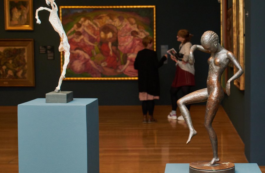 Im Raum stehen zwei sich zugewandte Frauen, an den Wänden hinter ihnen hängen mehrere große, mit vergoldeten Rahmen versehene Gemälde. Den Vordergrund des Fotos dominieren zwei dynamisch wirkende Skulpturen tanzender Körper.