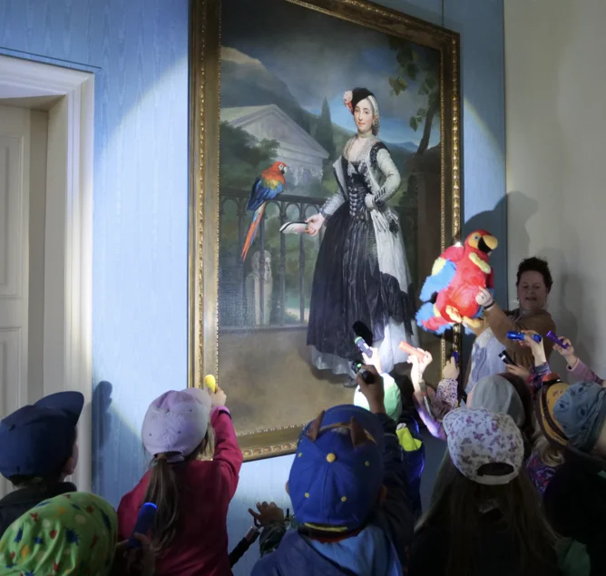 Eine Kindergruppe im Museum leuchtet ein großes Gemälde mit Taschenlampen an, daneben eine Frau mit einem Plüschpapagei.