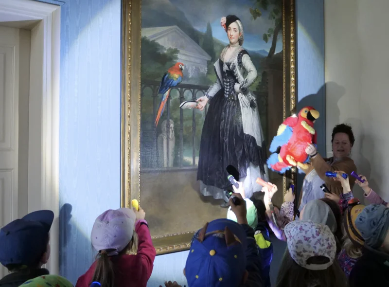 Eine Kindergruppe im Museum leuchtet ein großes Gemälde mit Taschenlampen an, daneben eine Frau mit einem Plüschpapagei.