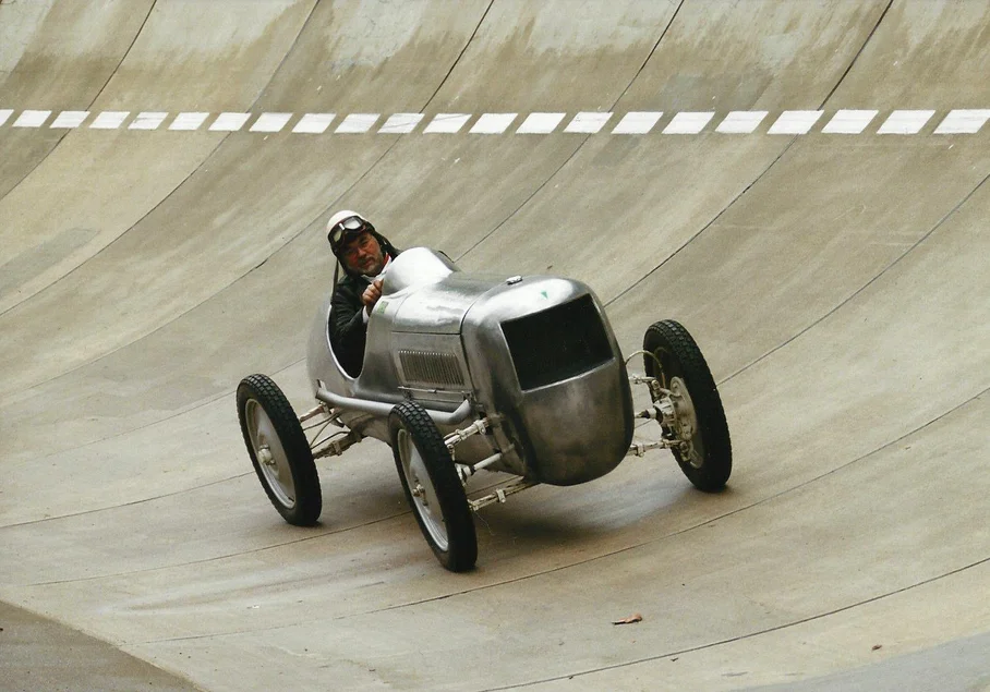 Frieder Bach im Fachsenfeld-Rennwagen Ein Mann fährt auf einer Rennstrecke einen historischen Rennwagen mit relativ kleiner Metallkarosse und relativ großen Rädern