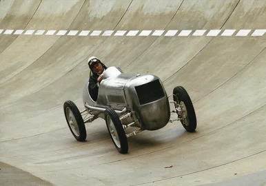Frieder Bach im Fachsenfeld-Rennwagen Ein Mann fährt auf einer Rennstrecke einen historischen Rennwagen mit relativ kleiner Metallkarosse und relativ großen Rädern