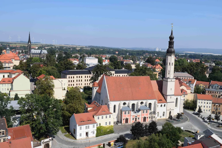 Farbfotografie von oben auf die Zittauer Altstadt, im Mittelpunkt eine große Kirche mit weißem Schiff, rotem Dach und grazilem, spitz zulaufendem Turm