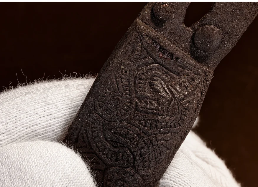 Verzierte Gürtelschließe Eine Hand in einem weißen Stoffhandschuh hält einen kleinen, schwarzen, mit Drachen verzierten archäologischen Fund.