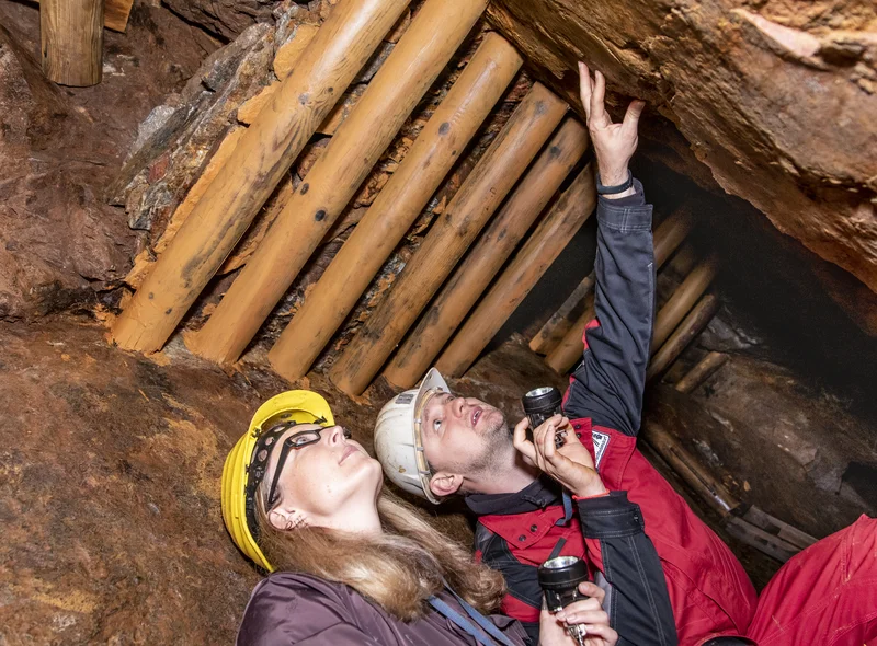 In einem Stolln, in dem die Decke durch Holzbalken verstärkt ist, hocken nebeneinander eine Besucherin mit Schutzkleidung (brauner Overall und Schutzhelm) und ein Mitarbeiter der Zinngrube (schwarz-rote Schutzjacke und Schutzhelm). Beide halten eine Taschenlampe in der Hand. Der Mitarbeiter deutet in die rechte obere Ecke des Bildes zu einer Steinwand, die Besucherin schaut in dieselbe Richtung.