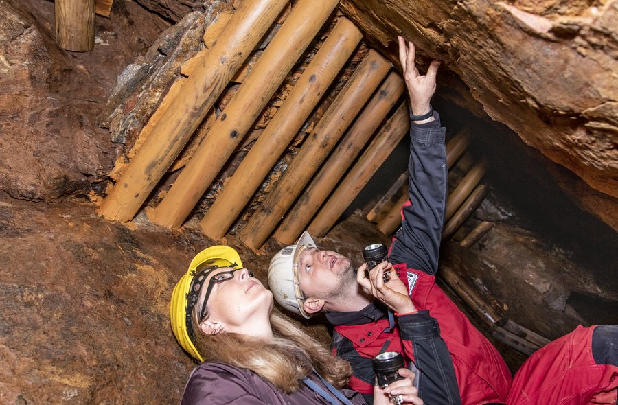 Historisch-bergbautechnologische Führung In einem Stolln, in dem die Decke durch Holzbalken verstärkt ist, hocken nebeneinander eine Besucherin mit Schutzkleidung (brauner Overall und Schutzhelm) und ein Mitarbeiter der Zinngrube (schwarz-rote Schutzjacke und Schutzhelm). Beide halten eine Taschenlampe in der Hand. Der Mitarbeiter deutet in die rechte obere Ecke des Bildes zu einer Steinwand, die Besucherin schaut in dieselbe Richtung.