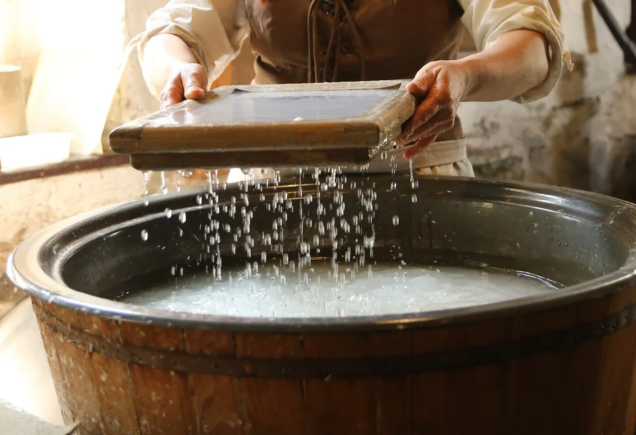 Der Prozess des Handschöpfens von Büttenpapier wird gezeigt, indem eine Person ein tropfendes Sieb über ein Fass hält.