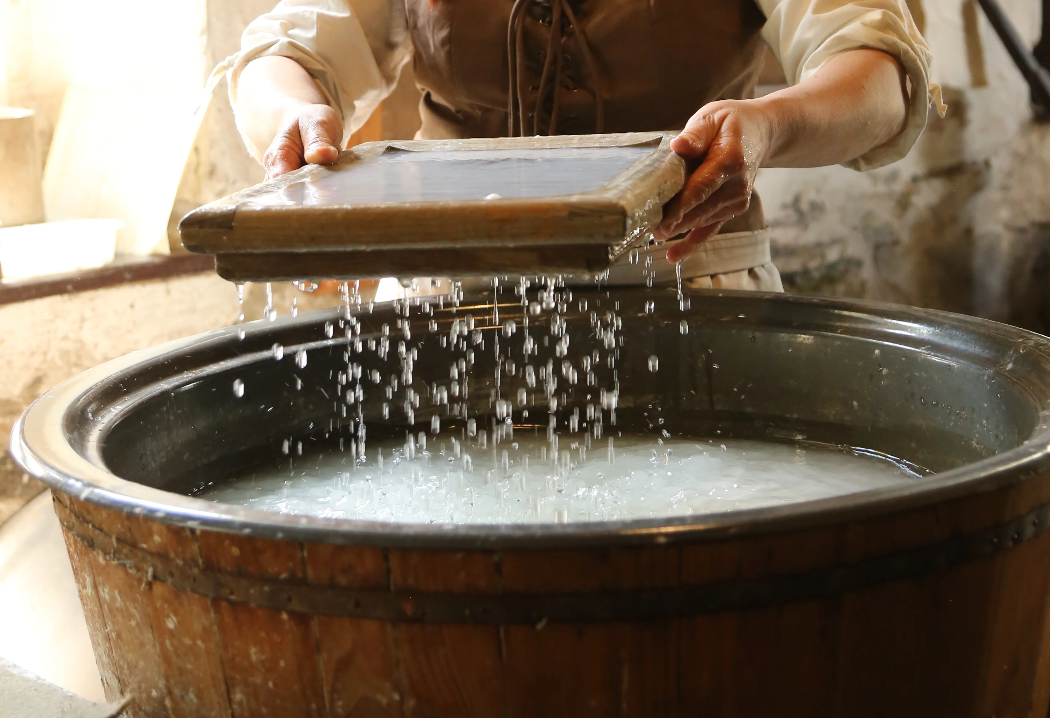 Papierschöpfen Der Prozess des Handschöpfens von Büttenpapier wird gezeigt, indem eine Person ein tropfendes Sieb über ein Fass hält.