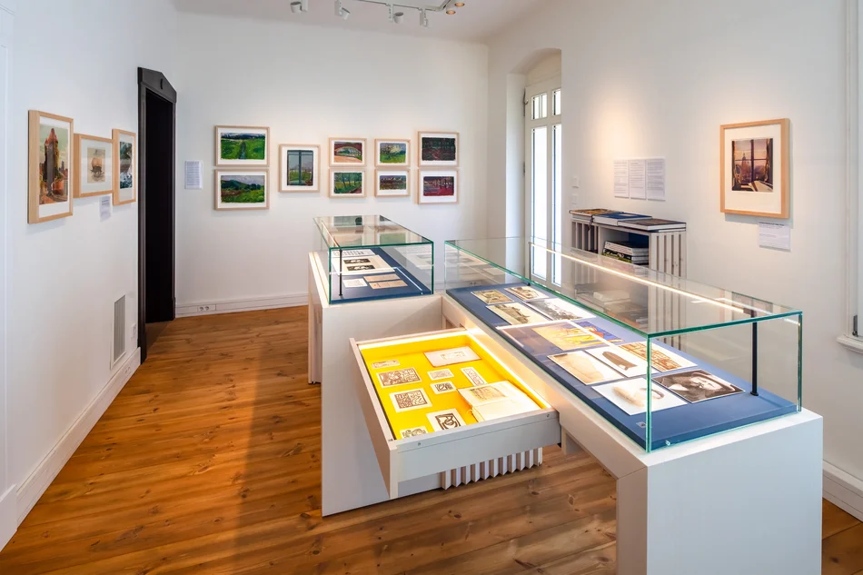Kleiner Ausstellungsraum mit Tischvitrinen, die Drucksachen beziehungsweise grafische Blätter enthalten, sowie gerahmten Bildern an den Wänden