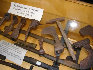 Bergmännisches Gezähe Neben einem Objektschild mit der Aufschrift "Werkzeuge der Bergleute. Fundorte: Alte Stollen im Auer Gebiet" befinden sich mehrere historische Eisenwerkzeuge.