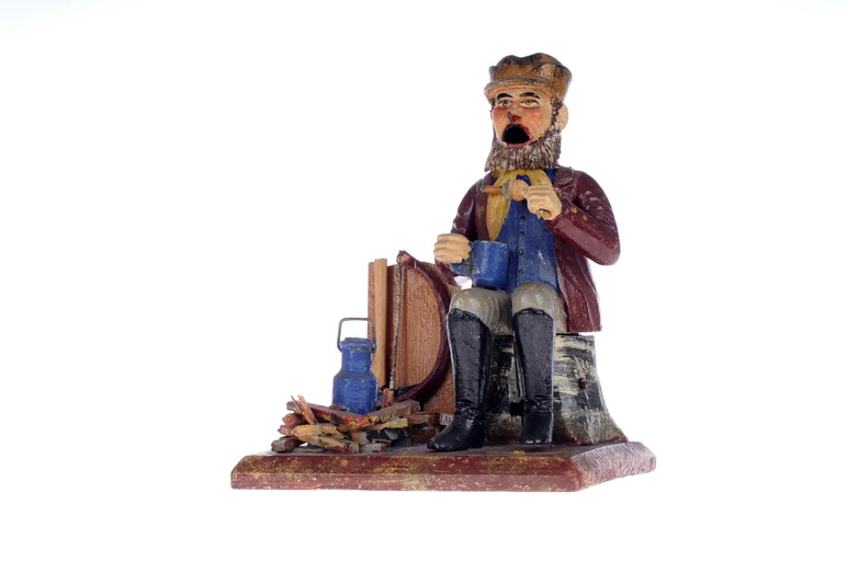 Das Foto zeigt ein original erzgebirgisches Räuchermannchen, geschnitzt aus Holz und bunt bemalt. Es ist die Figur eines historisch gekleideten Waldarbeiters, der, auf einem Baumstumpf sitzend, eine Brotzeit einnimmt und in der rechten Hand eine große Tasse hält.