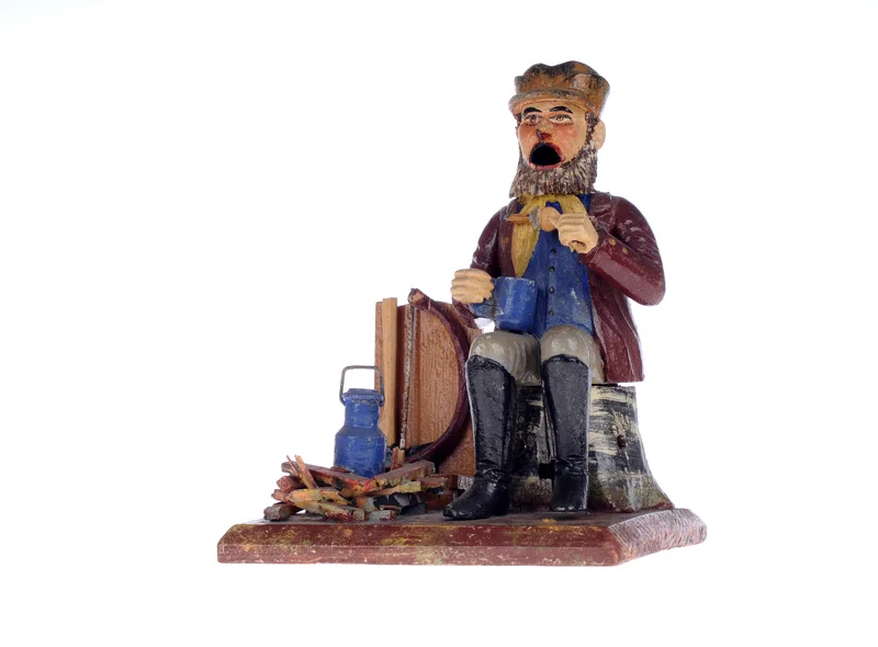 Das Foto zeigt ein original erzgebirgisches Räuchermannchen, geschnitzt aus Holz und bunt bemalt. Es ist die Figur eines historisch gekleideten Waldarbeiters, der, auf einem Baumstumpf sitzend, eine Brotzeit einnimmt und in der rechten Hand eine große Tasse hält.