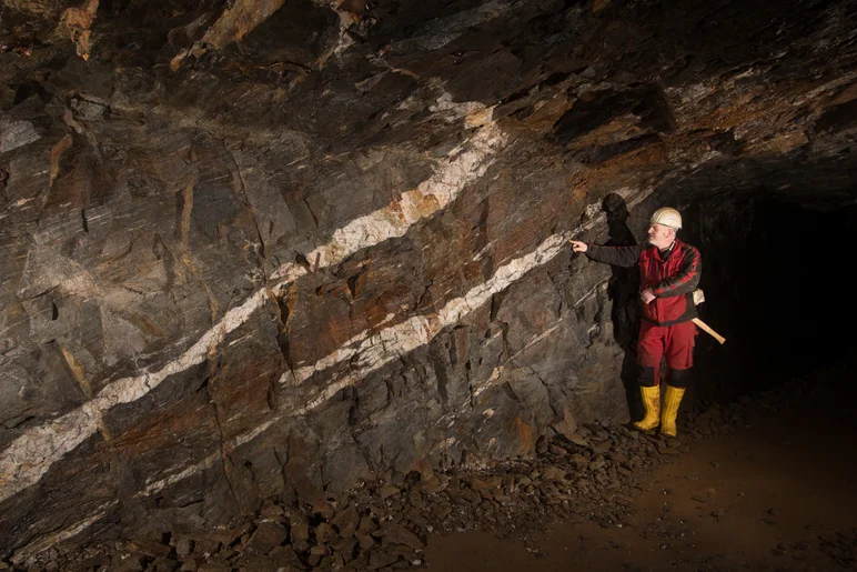 In einem Stollen steht ein Mann in Schutzkleidung (Jacke, Hose, Stiefel, Schutzhelm) vor einer Steinwand. In dieser Wand sind zwei dicke, quer von links unten nach rechts oben verlaufende, weiße Erzadern zu sehen, die etwa die Breite einer Hand eines Erwachsenen haben. Um diese weißen Erzadern herum befindet sich dunkelgraues Gestein. Der Mann zeigt auf die obere der beiden Erzadern.