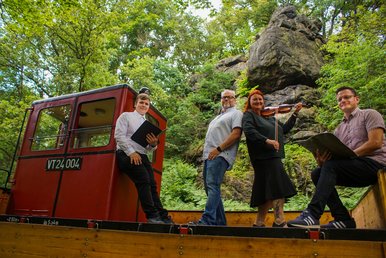 Sommerreise mit Musik und Poesie Drei Männer und eine Geige spielende Frau stehen beziehungsweise sitzen auf einem offenen historischen Güterwaggon.