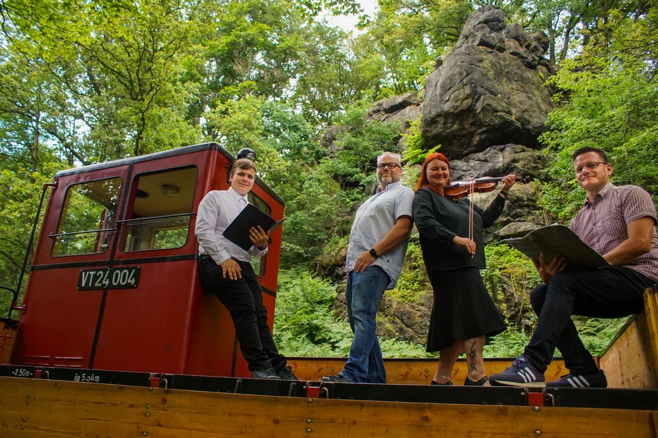Sommerreise mit Musik und Poesie Musiker und Interpreten stehen auf einem Museumsbahnzug in grüner Landschaft, im Hintergrund ragt ein Felsen auf.