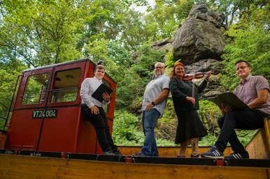 Sommerreise mit Musik und Poesie Musiker und Interpreten stehen auf einem Museumsbahnzug in grüner Landschaft, im Hintergrund ragt ein Felsen auf.