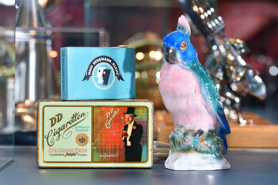 Das Foto zeigt einen Rauchverzehrer in Form eines Papageien und zwei Zigarettenschachteln der Marken „Haus Bergmann“ und „DD Cigaretten“.
