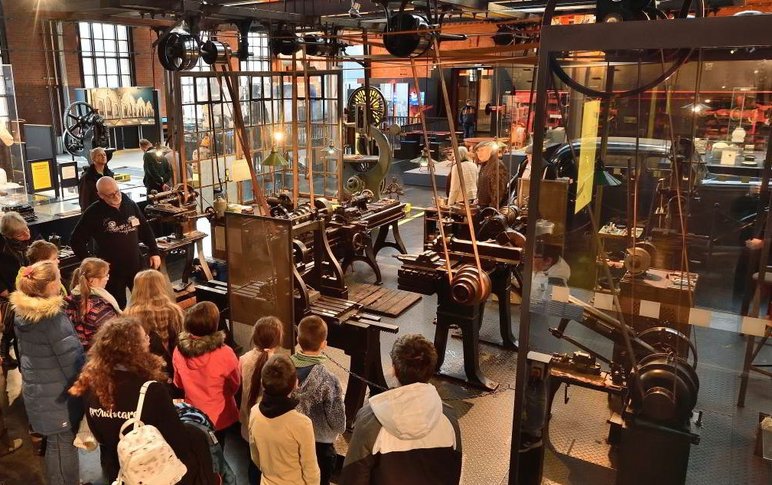 Eine Schulklasse und ein erwachsener Museumsmitarbeiter stehen vor der in der Dauerausstellung inszenierten Schlosserwerkstatt mit ihren Originalmaschinen und -werkzeugen.