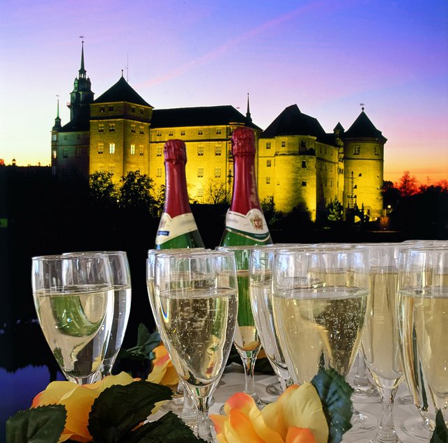 Collage zweier Fotos. Oben: ein in der Abenddämmerung illuminiertes Schloss. Unten: Zwei Sektflaschen mit mehreren Sektgläsern.