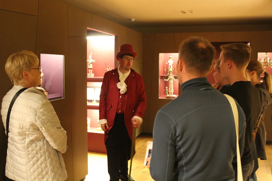 In einem mit Vitrinen bestückten Ausstellungsraum hört eine Besuchergruppe einem Museumsführer in historischem Kostüm zu. In den beleuchteten Vitrinen sind Objekte erzgebirgischer Volkskunst erkennbar.
