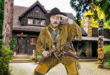 Old Shatterhand wartet auf Sie! Ein Mann in Cowboy-Kostüm und mit einem Gewehr in der Hand steht spähend vor einem großen Blockhaus.