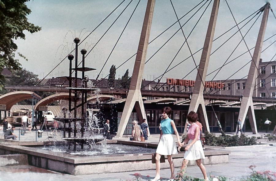 Farbfotografie eines Brunnens vor einem Flachbau im Stil der Ostmoderne, dessen Dach von Stahlseilen an schräg aufragenden Betonpfeilern gehalten wird. Im Vordergrund zwei sommerlich gekleidete Frauen im Siebziger-Jahre-Stil.