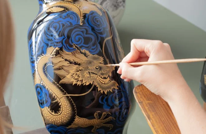 Eine Hand malt mit einem schmalen Pinsel ein goldenes Drachen-Motiv auf eine dunkelblaue Porzellanvase.