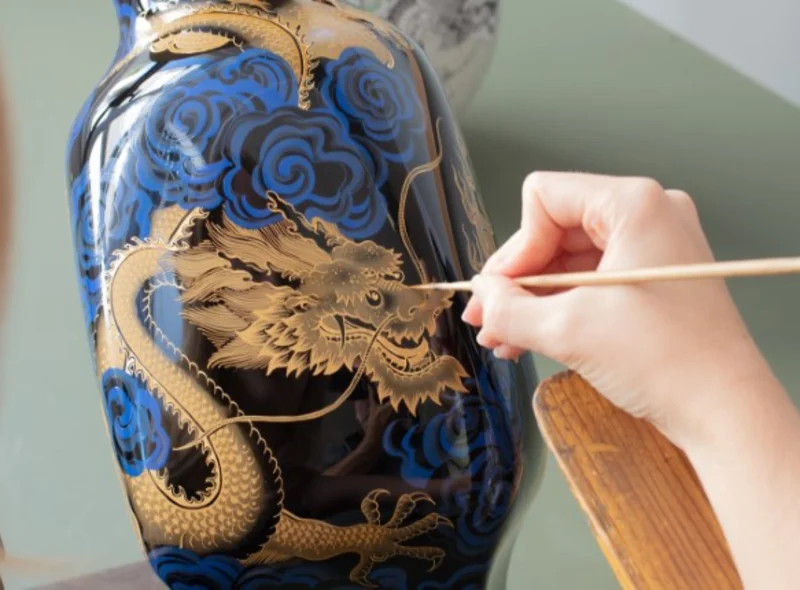 Eine Hand malt mit einem schmalen Pinsel ein goldenes Drachen-Motiv auf eine dunkelblaue Porzellanvase.