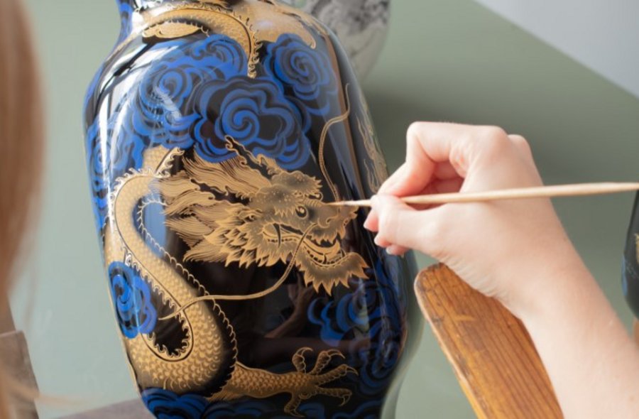 Drachenführung Eine Hand malt mit einem schmalen Pinsel ein goldenes Drachen-Motiv auf eine dunkelblaue Porzellanvase.