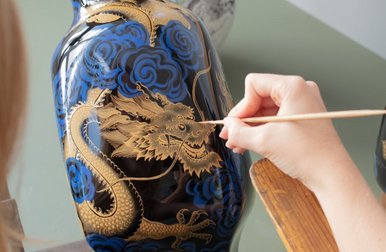 Drachenführung Eine Hand malt mit einem schmalen Pinsel ein goldenes Drachen-Motiv auf eine dunkelblaue Porzellanvase.