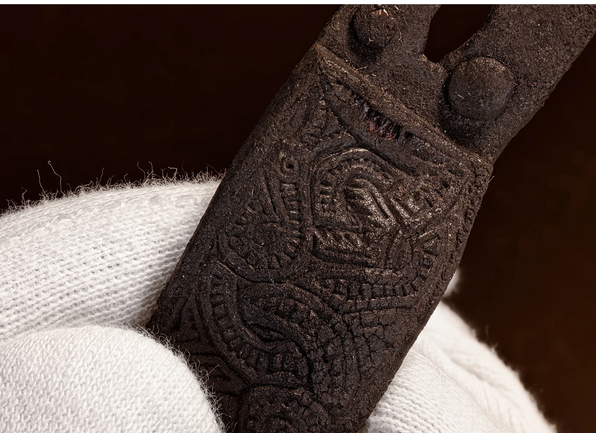 Verzierte Gürtelschließe Eine Hand in einem weißen Stoffhandschuh hält einen kleinen, schwarzen, mit Drachen verzierten archäologischen Fund.