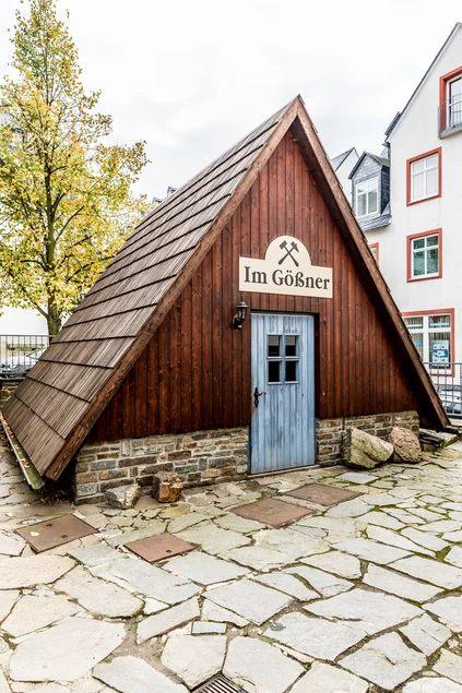 Auf einem historischen Innenhof steht ein kleines Holzgebäude mit einem bis auf den Boden reichenden Spitzdach und einem Natursteinsockel.