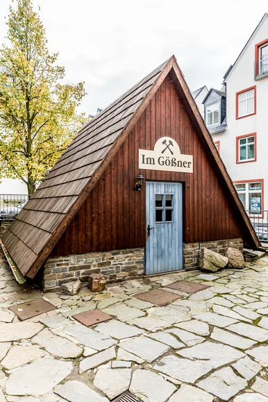 Auf einem historischen Innenhof steht ein kleines Holzgebäude mit einem bis auf den Boden reichenden Spitzdach und einem Natursteinsockel.