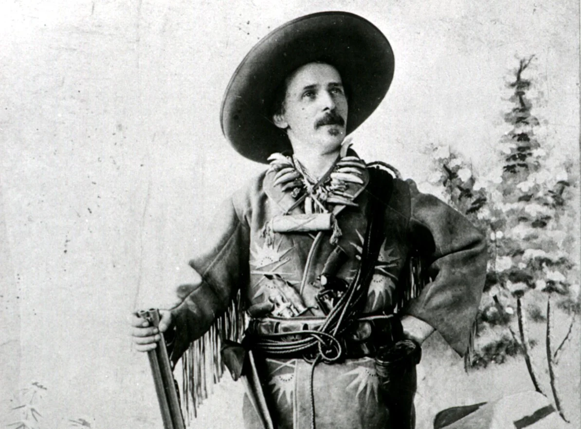 Karl May posiert im Old-Shatterhand-Kostüm vor der Kamera mit Gewehr, Lederjacke und Hut.